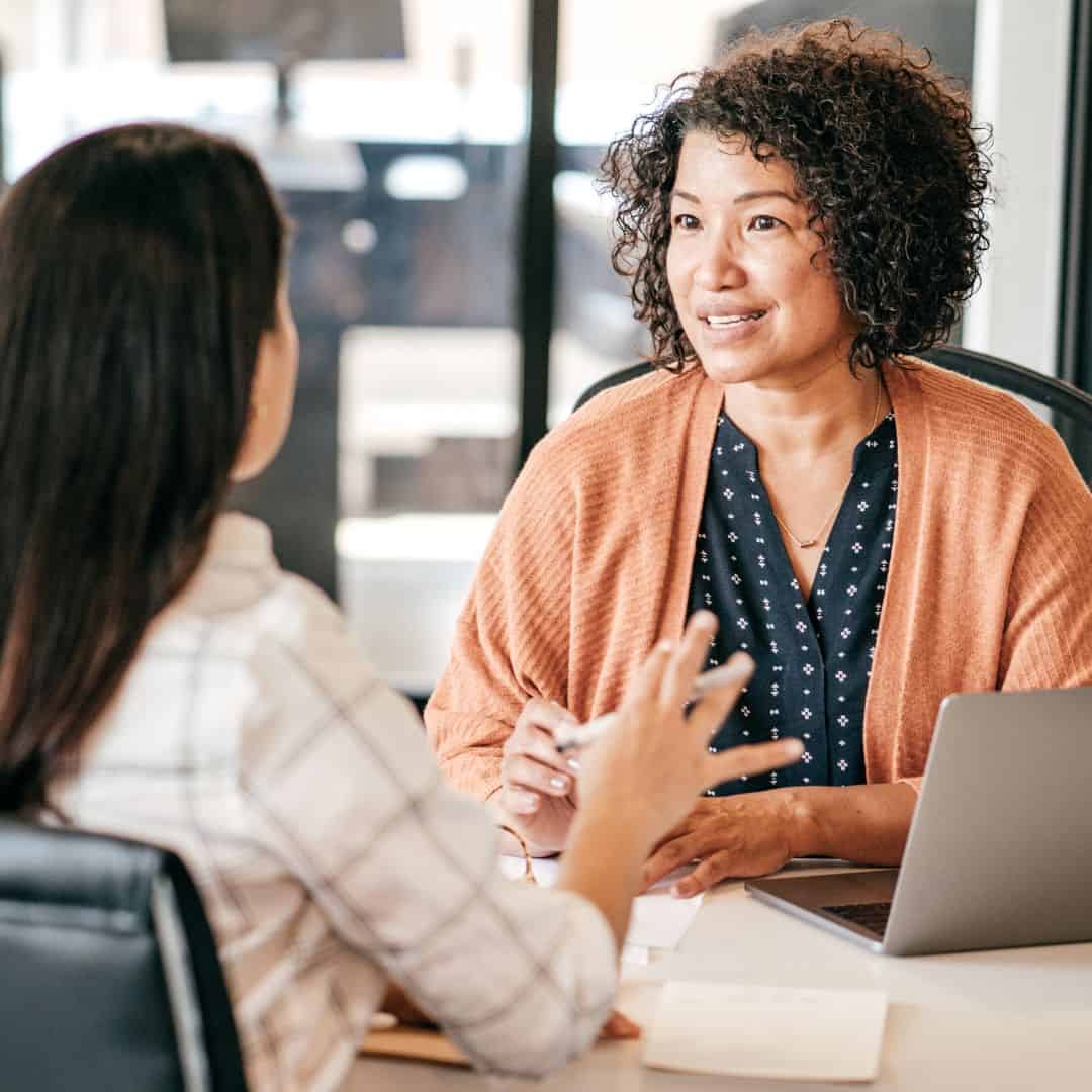 Two educators discussing interview preparation across a table with a laptop